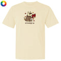 A cream-colored short-sleeve t-shirt with a cartoon graphic of a dog wearing sunglasses and reading a red book, with the text 'all booked up' below. A small multicolor color wheel icon is in the top-left corner.