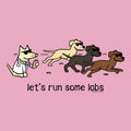 A pink graphic print featuring a cartoon dog in a white lab coat running behind three other dogs wearing sunglasses, with the text 'let's run some labs' below. Small text at the bottom right reads 'WE'VE BOTH OWNED DOGNAME'.