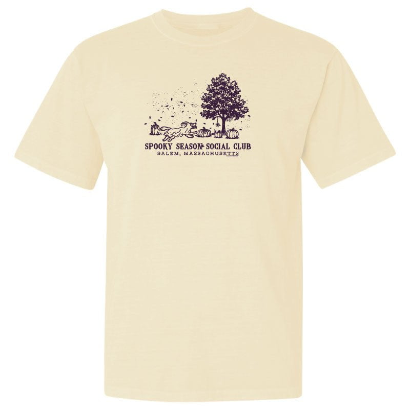A cream-colored short-sleeve t-shirt with a dark purple graphic on the chest depicting a tree, pumpkins, and the text 'Spooky Season Social Club' and 'Salmon, Massachusetts'.
