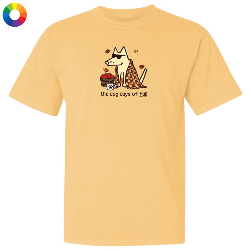 A pale yellow short-sleeve t-shirt with a graphic of a dog wearing sunglasses and a plaid scarf, next to a basket of red apples, with the text 'the dog days of fall' printed below. A small multicolor color wheel icon is in the top-left corner.